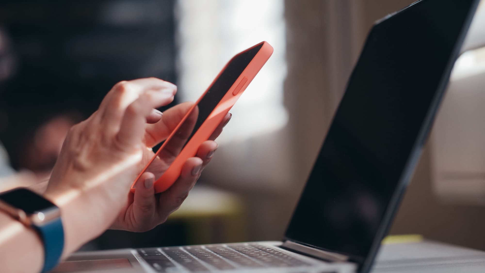 A hand holding a smartphone with an orange case, in front of a laptop