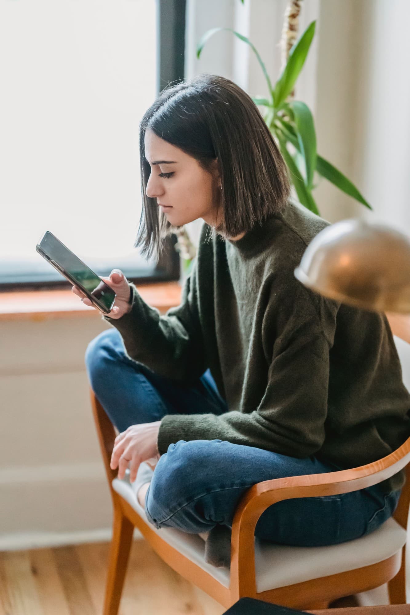 Une jeune femme assise dans un fauteuil consulte son téléphone près d’une fenêtre